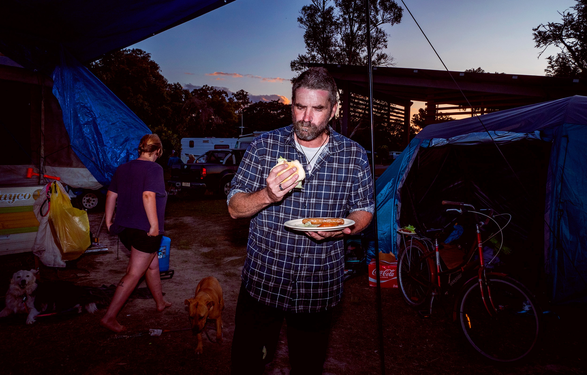 A man eats a burger surrounded by tents as evening falls.