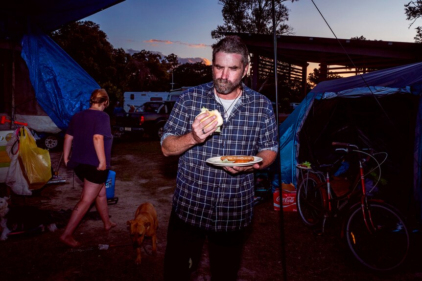 A man eats a burger surrounded by tents as evening falls.