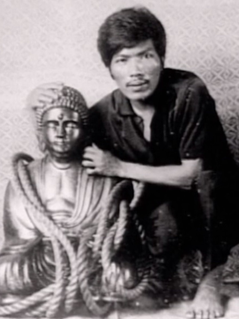 A black and white photo of a man with his hands wrapped around a shiny buddha statue, covered in a long rope