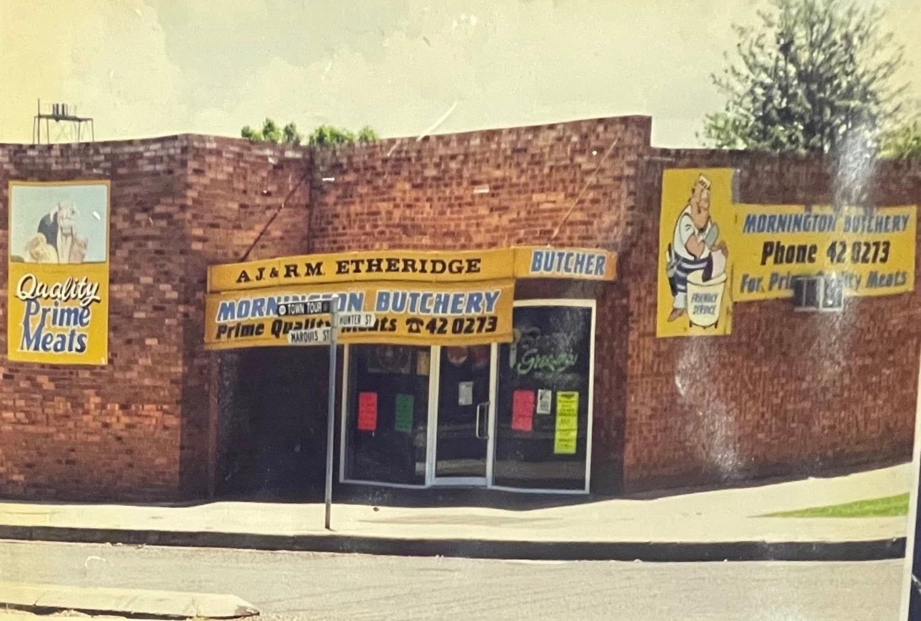 লাল ইট মর্নিংটন কসাই দোকান সামনে পুরানো ছবি. দোকানটি রঙিন সাইনবোর্ড এবং কার্টুন কসাই চরিত্র দিয়ে সজ্জিত করা হয়েছে।