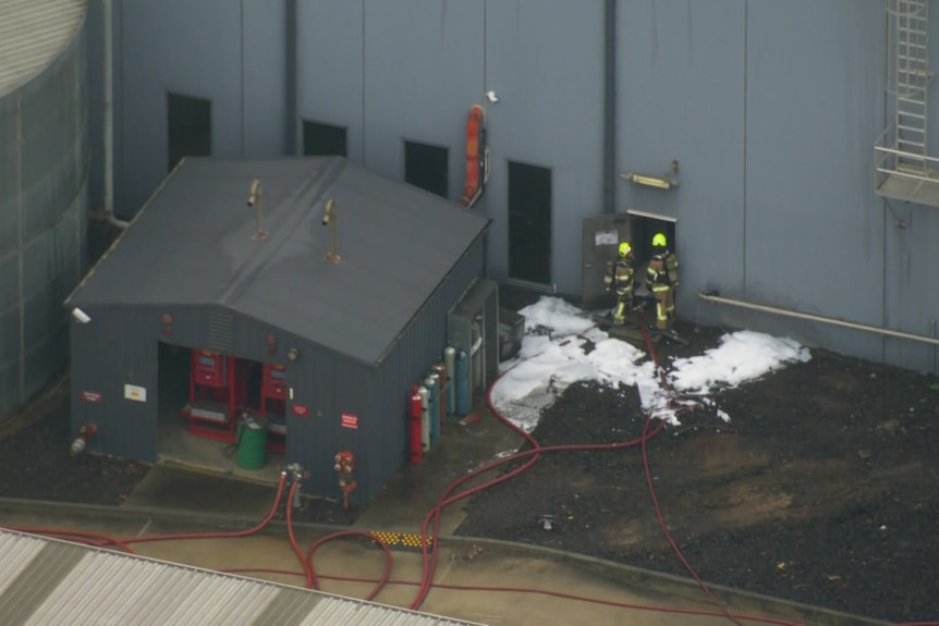Two firefighters stand at the open door of a factory where firefighter foam covers the ground.