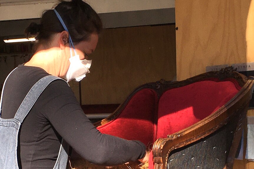 Susan is fixing an old red chair. The fibres from inside the chair are exposed and Susan is wearing a breathing mask.