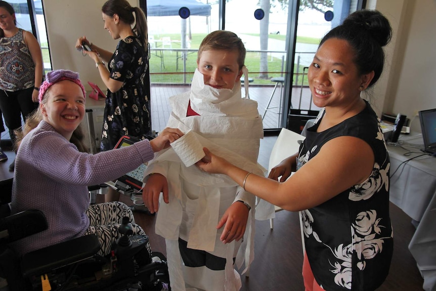 A woman in a wheelchair wraps a boy up in toilet paper like a mummy