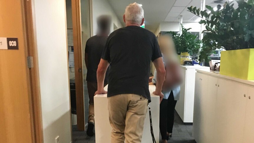 A man whose back is towards the camera walks through an office. Two other people in are blocked out.