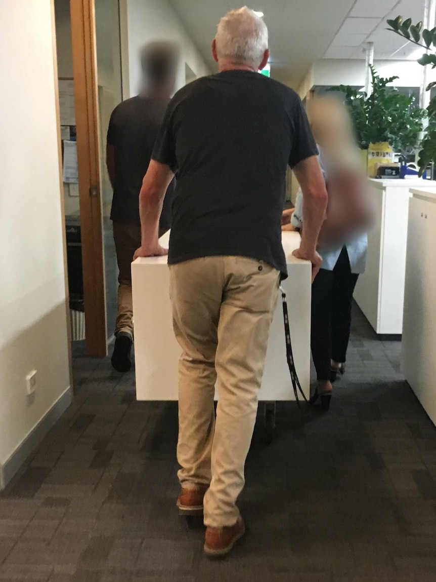 A man whose back is towards the camera walks through an office. Two other people in are blocked out.