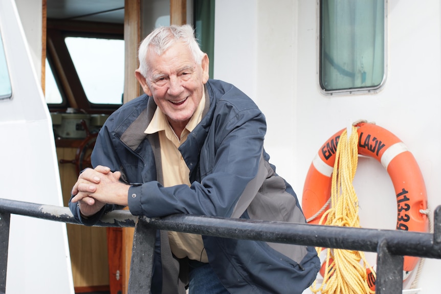 Man smiling, looking at camera, leaning on rail of boat, lifebuoy in background, 