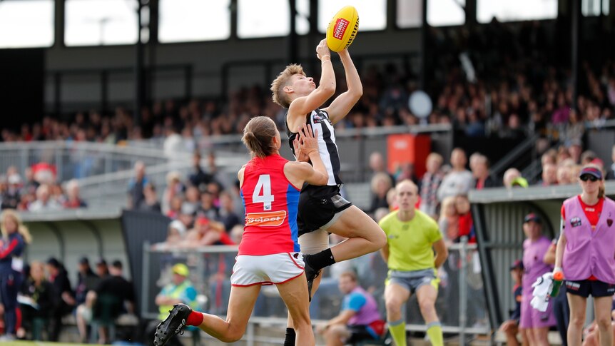 A Collingwood AFLW player leaps with hands extended to take a mark as a Melbourne player pushes her in the side.