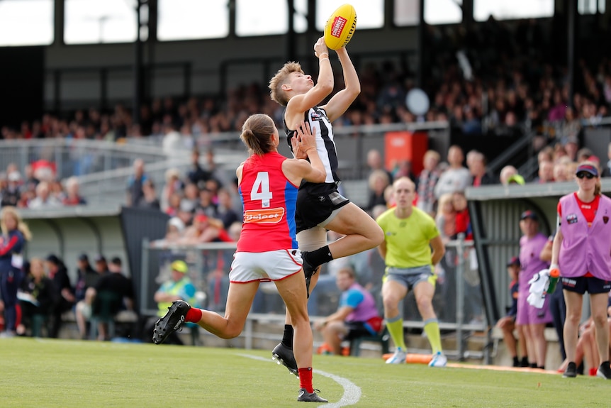 A Collingwood AFLW player leaps with hands extended to take a mark as a Melbourne player pushes her in the side.