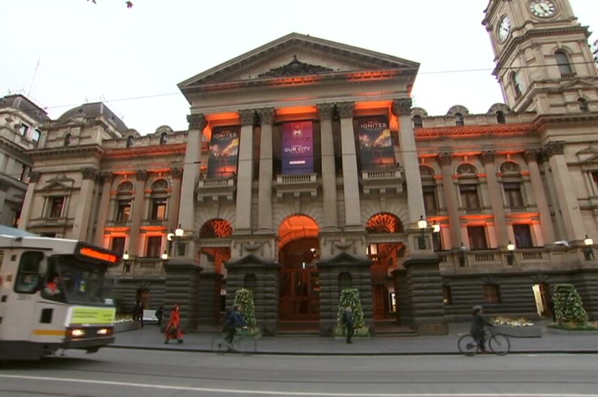 Melbourne Town Hall is lit with orange lights in its arches against a grey sky on Collins Street as a tram moves past.