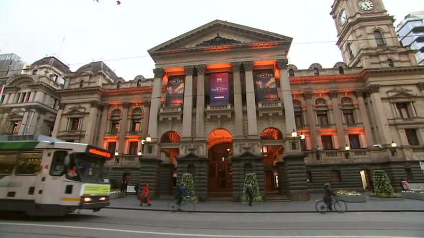 Melbourne Town Hall is lit with orange lights in its arches against a grey sky on Collins Street as a tram moves past.
