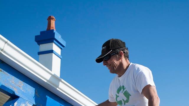 A man in shorts, t-shirt, sunglasses and cap stands on a ladder painting the exterior timber lintels of a building blue.