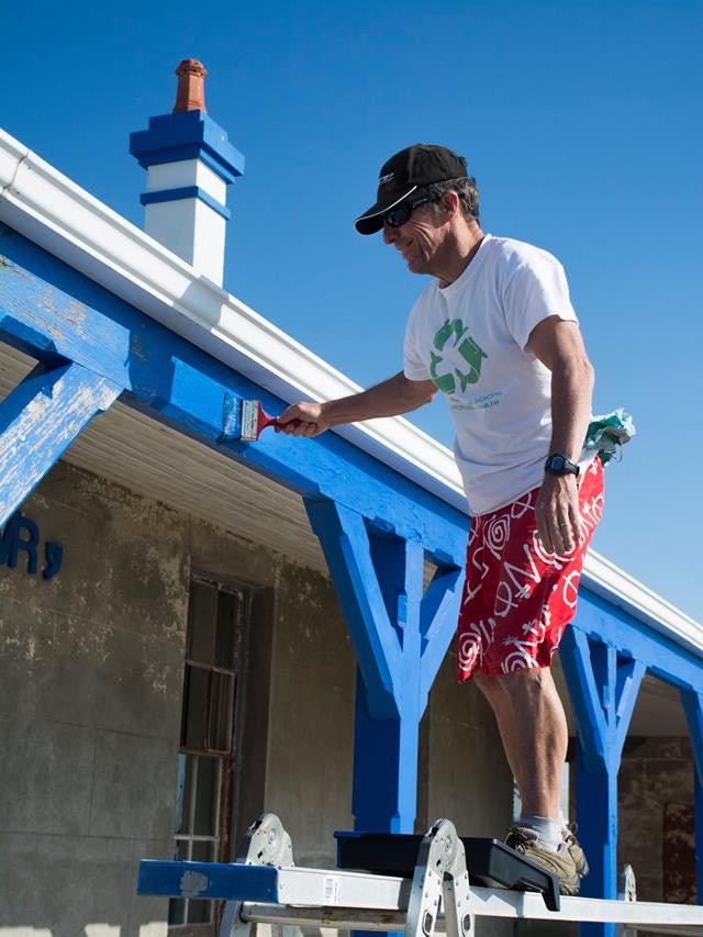 A man in shorts, t-shirt, sunglasses and cap stands on a ladder painting the exterior timber lintels of a building blue.