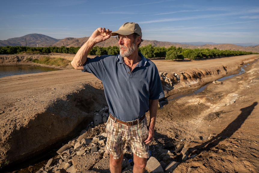 A man in shorts, polo shirt and cap stands on a dusty plain looking out at the horizon