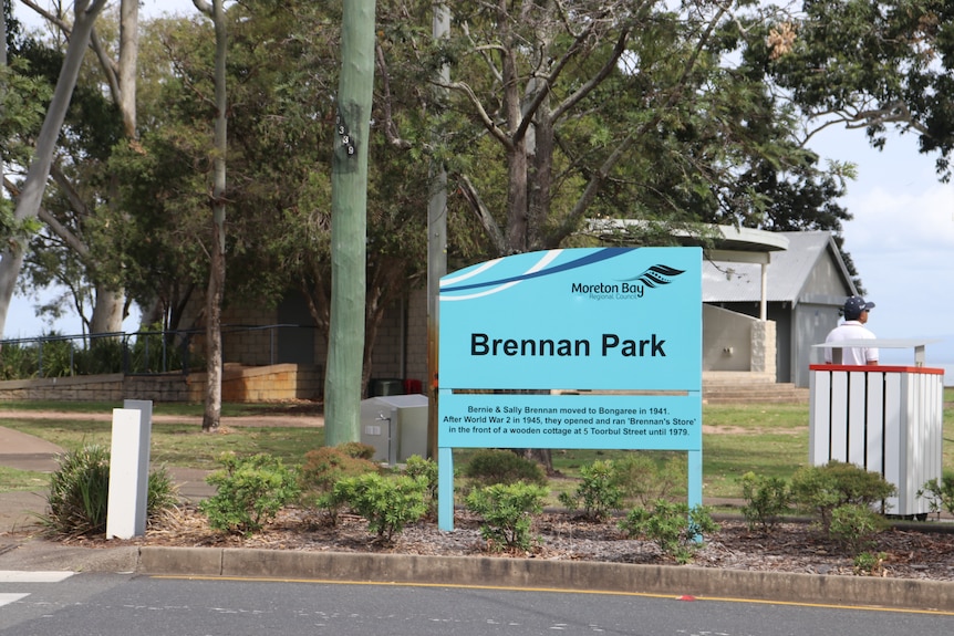 A light blue sign that reads 'Brennan Park' sits in a roadside garden bed.