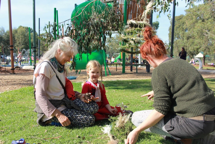 An older woman, a young girl and a young woman sit on lawn next to a playground.