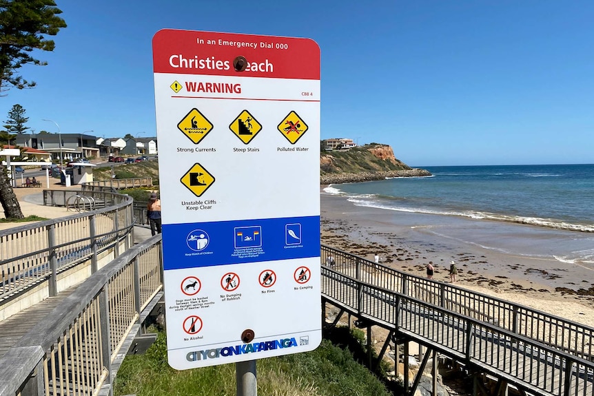 A sign issues several warnings near a ramp down a beach with cliffs in the background.