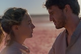 A white blonde woman and a white brunette man, both in their 30s, stand intimately looking at each other in a desert.
