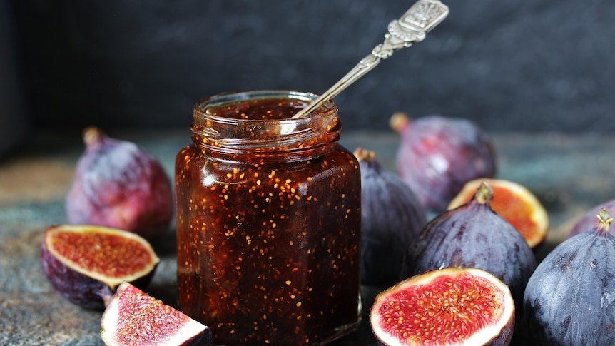 Several sliced-open figs scattered around a jar of fig jam with a spoon in it.