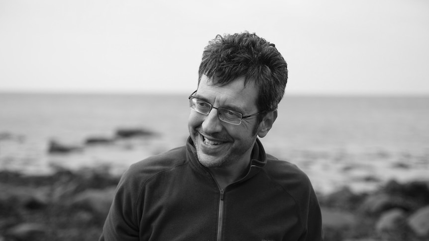 You see a black and white portrait of George Monbiot smiling and looking off camera on a rocky beach.