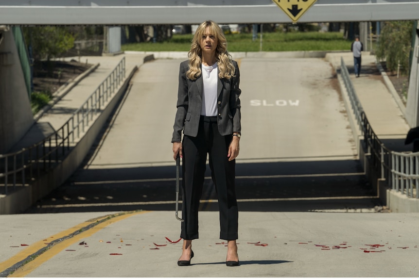 Exterior daylight shot of young woman with long light blonde hair in suit, standing in middle of empty road holding tire iron.