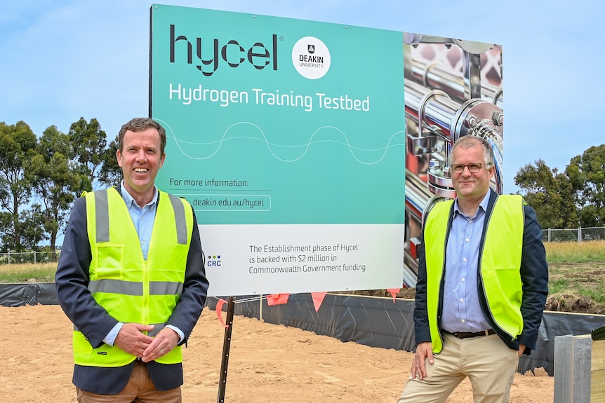 Two men wearing high vis jackets looking at the camera in front of a sign and construction site.