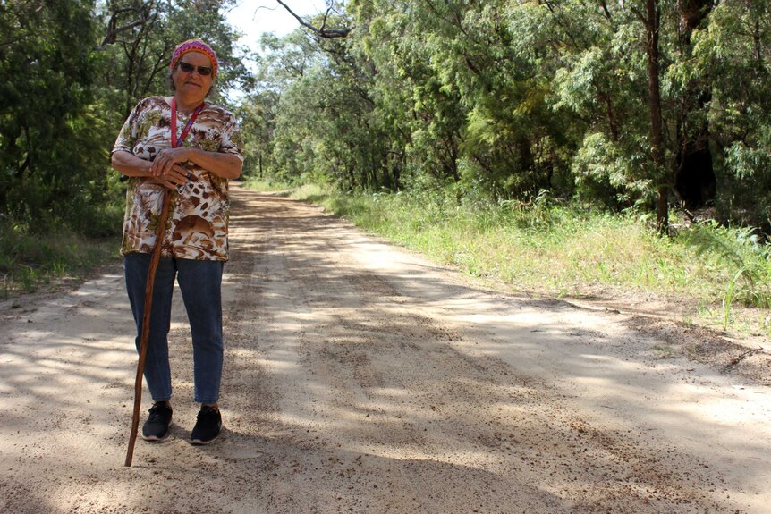 A woman stands in the centre of a gravel road holding a walking stick.