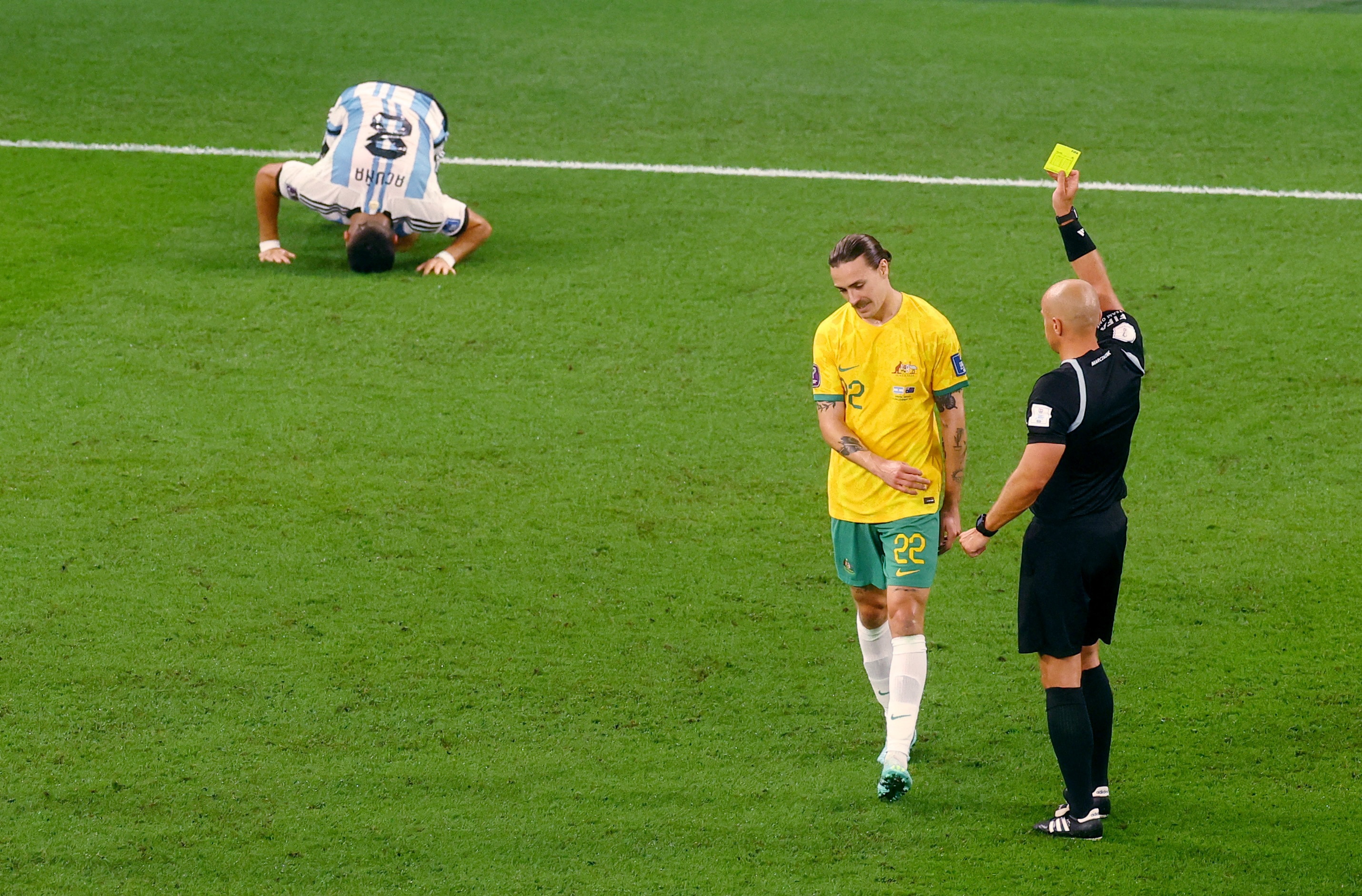 A referee holds up a yellow card for Australian midfielder Jackson Irvine as an Argentinian player hunches on the pitch.