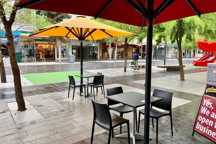 empty tables and chairs sit below an umbrella in a shopping strip on a wet day