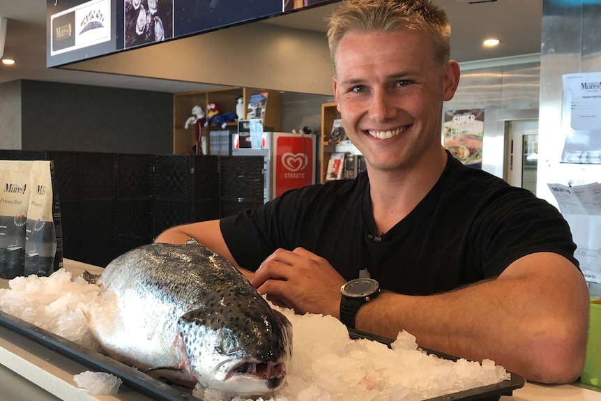 A man smiles behind the counter of a seafood store. A fish is on display in front of him