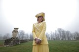 A melancholy-looking 30-something woman with a blonde bob, wearing a yellow blazer, skirt and hat, stands in the countryside