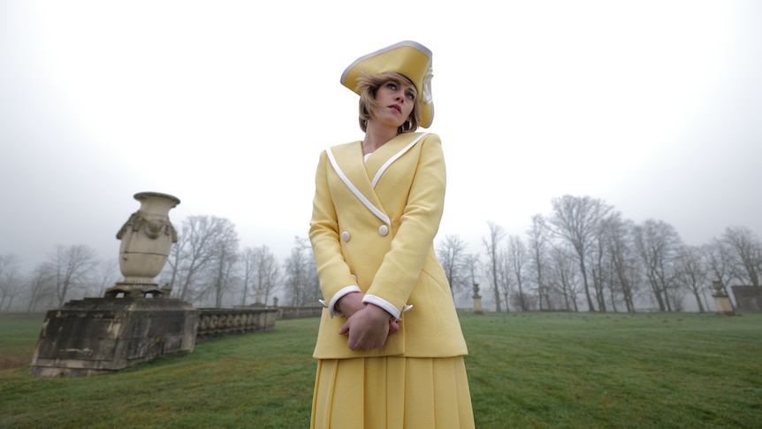 A melancholy-looking 30-something woman with a blonde bob, wearing a yellow blazer, skirt and hat, stands in the countryside