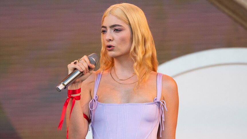 A blonde-haired Lorde in a lavendar top performing live with a silver mic in her hand