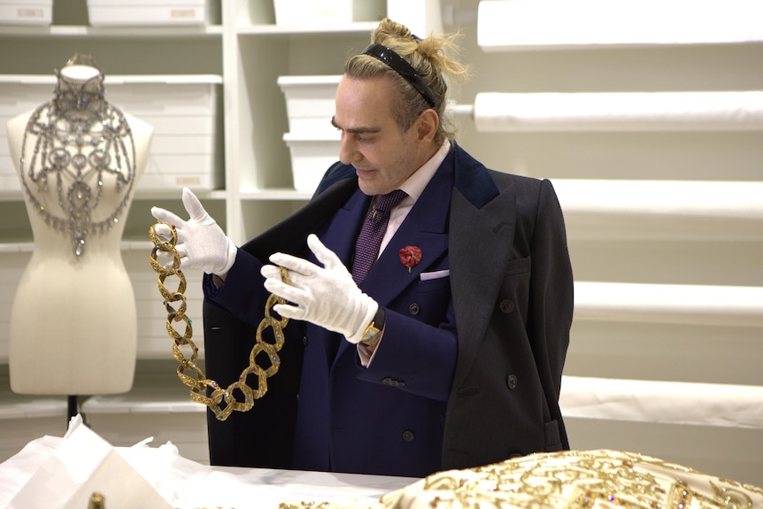 The designer John Galliano, in his 60s, in a navy suit and grey overcoat, holding a large piece of jewellery 