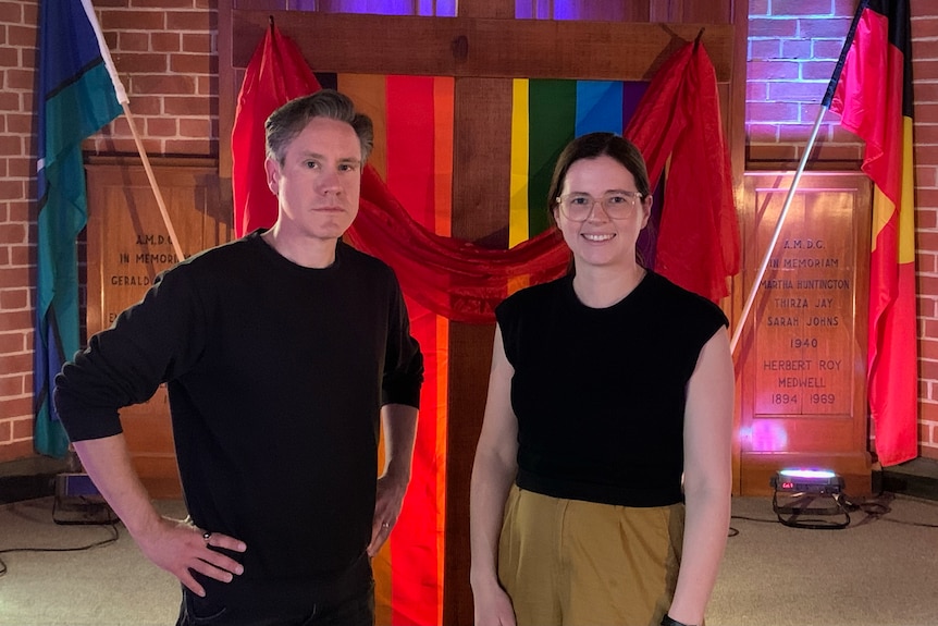 Anthony Castle and Rosalie Dow Schmidt standing in a church, with a rainbow flag in background.