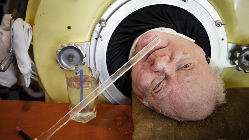 Polio survivor Paul lying down in an iron lung with a glass of water next to his head