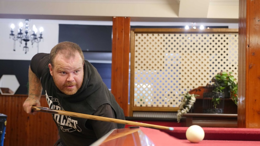A man lines up a shot at a pool table holding the cue in his right hand, left arm pointing to the ground.