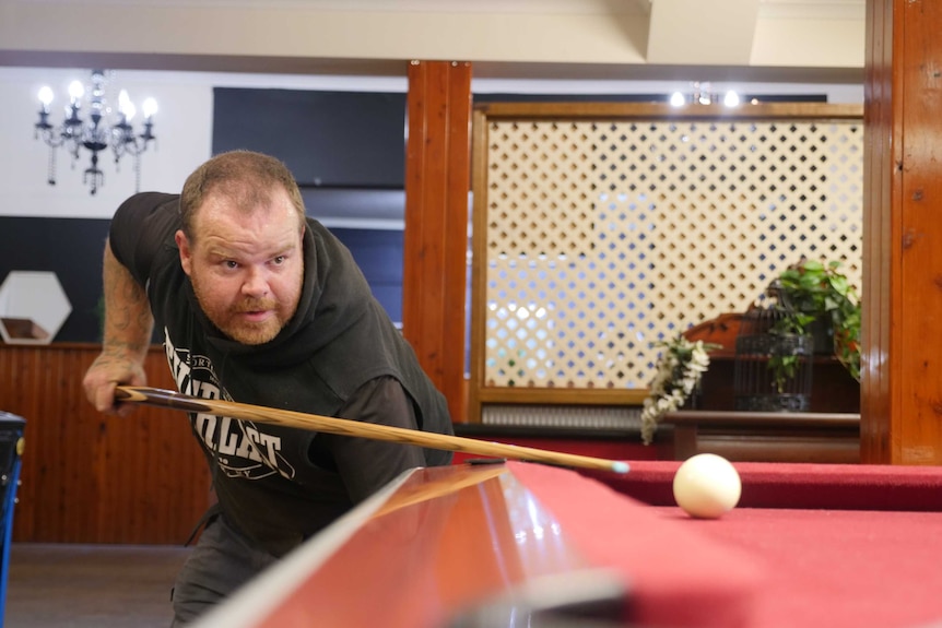 A man lines up a shot at a pool table holding the cue in his right hand, left arm pointing to the ground.