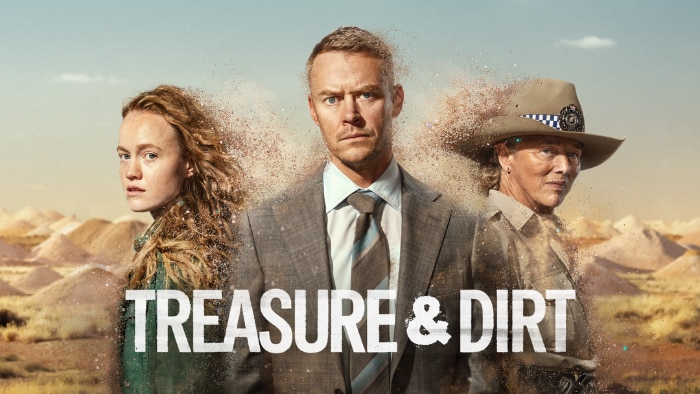 A woman with curly hair, a man in a suit, and a woman wearing a hat stand before a dusty landscape with mounds of dirt.