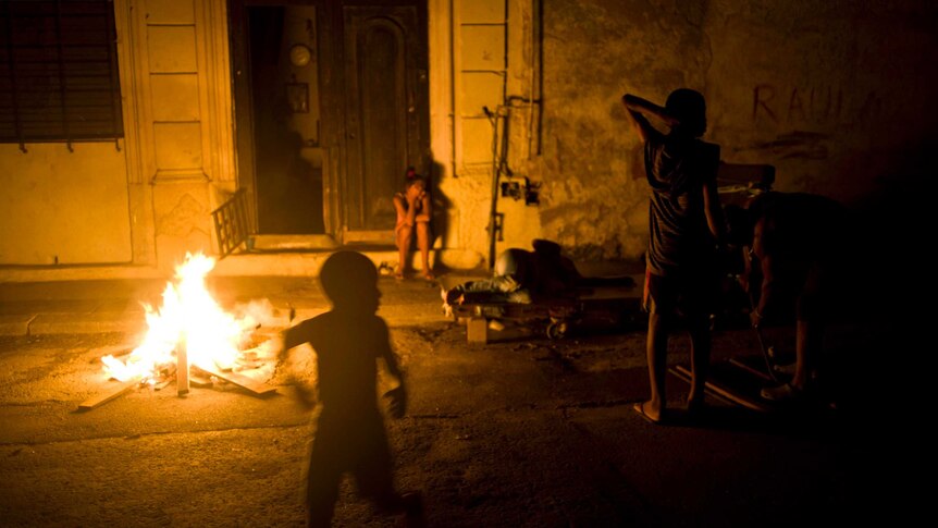 A few people, including children, are gathered near a fire outside a property at night.