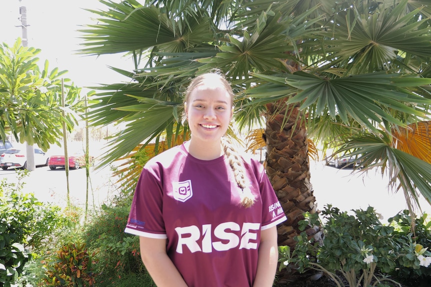 A young woman wearing a maroon shirt stands in front of trees smiling