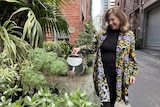 A lady in a colourful coat smiles as she waters plants in a laneway