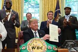 Donald Trump sitting at a desk holds up a piece of paper surrounded by people including Sylvester Stallone and Lennox Lewis.