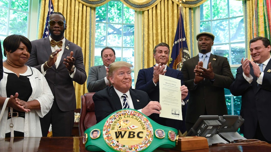 Donald Trump sitting at a desk holds up a piece of paper surrounded by people including Sylvester Stallone and Lennox Lewis.