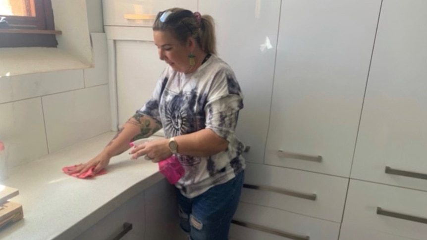 A woman uses pink cleaning products to clean a white kitchen.