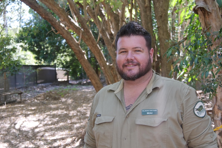 A smiling man in a khaki  shirt stands smiling in a wildlife park.