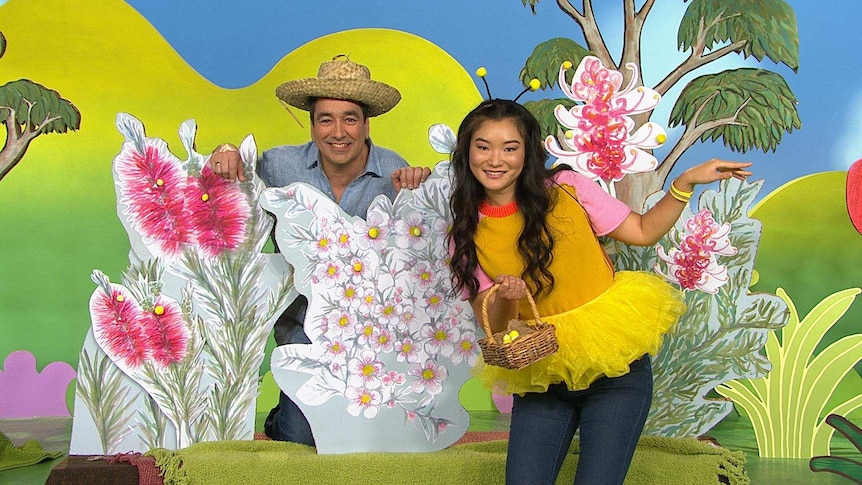 Alex wearing a hat, Michelle wearing a bee costume, on the Play School set surrounded by plants