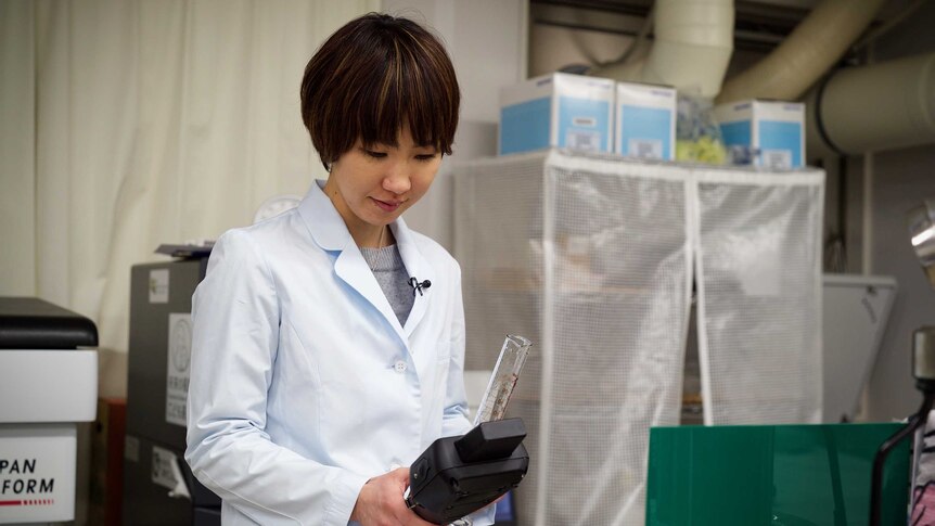 A Japanese woman in a lab coat looks at a radiation testing machine