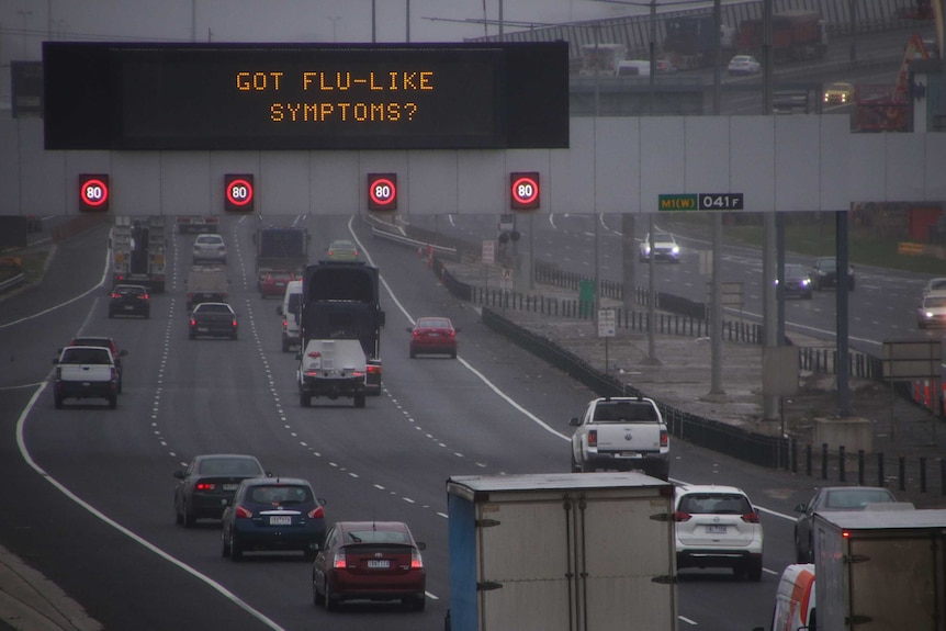 A large digital sign reads "got flu-like symptoms?" over a freeway on a cloudy and grey Melbourne day.