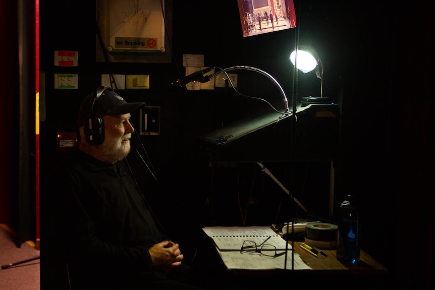 A man with gray beard sits at darkened side stage desk, monitoring scripts and screens.