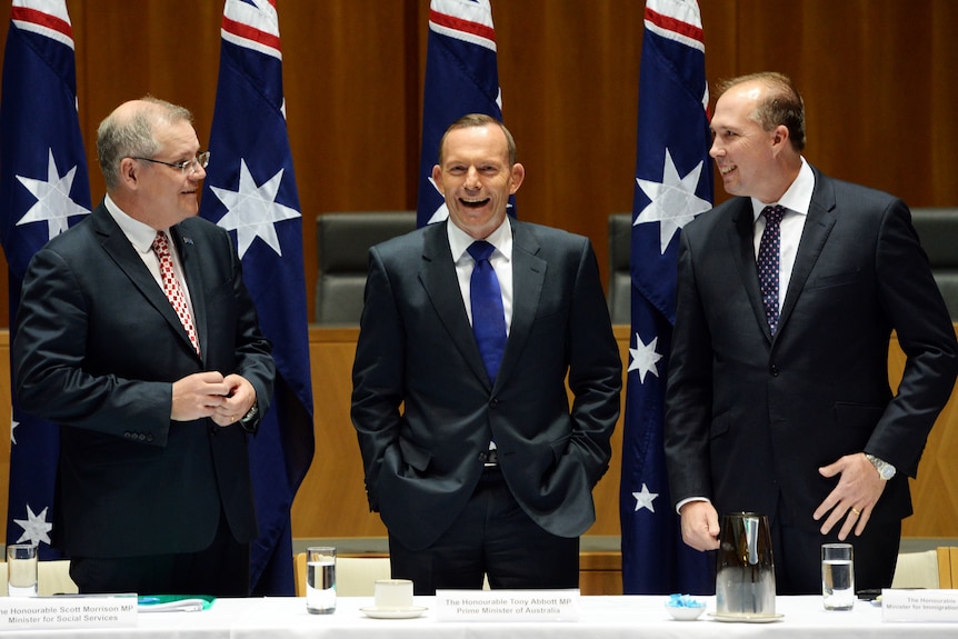 three men standing behind a desk talking and smiling